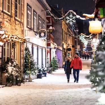 A couple takes a walk in the evening during the holiday season on rue du Petit-Champlain, covered with snow and decorated with illuminated trees.