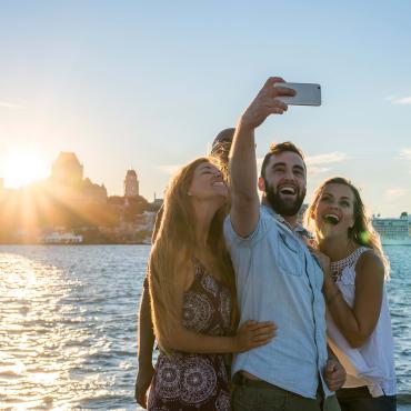 Des amis se prennent en photo au coucher du soleil, près du fleuve à Lévis avec un panorama du Vieux-Québec à l'arrière.